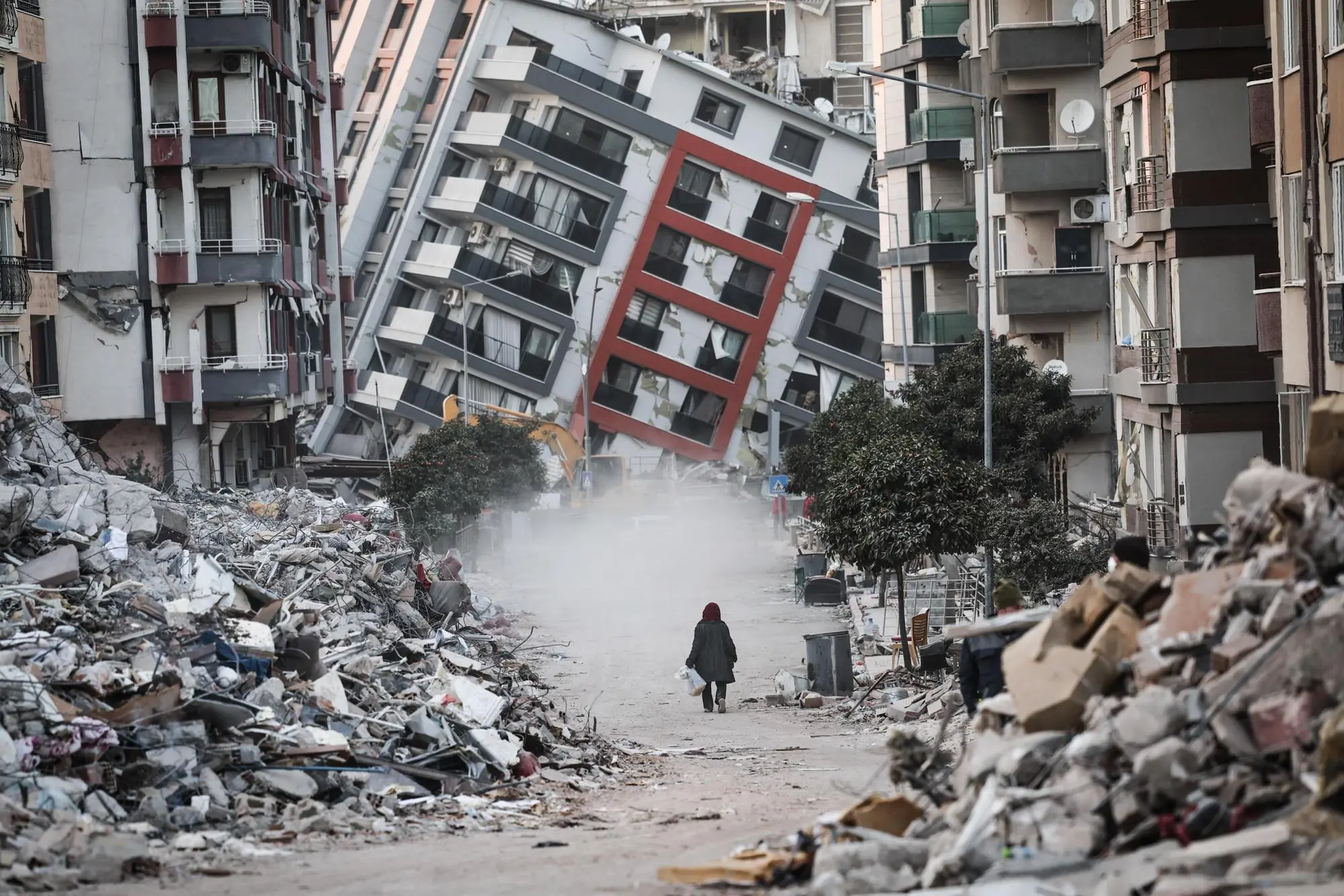 epaselect epa10473572 A woman walks in front of a collapsed building while demolition teams work after a powerful earthquake in Hatay, Turkey, 17 February 2023. Almost 44,000 people have died and thousands more are injured after two major earthquakes struck southern Turkey and northern Syria on 06 February. Authorities fear the death toll will keep climbing as rescuers look for survivors across the region. EPA/SEDAT SUNA