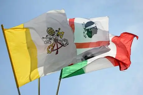 Una bandiera vaticana bianca e gialla con l'effigie papale, insieme a una bandiera sarda e una italiana